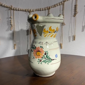 May include: A white ceramic vase with a frog-like design, featuring yellow and orange floral accents. The vase has a triangular opening and the text "VI.SE.PUOI" is written on the side. The vase is on a wooden table.