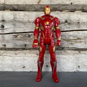 May include: A red and gold Iron Man action figure stands against a wooden background. The figure has a detailed design with gold accents on the arms, legs, and helmet. The figure is a collectible toy.