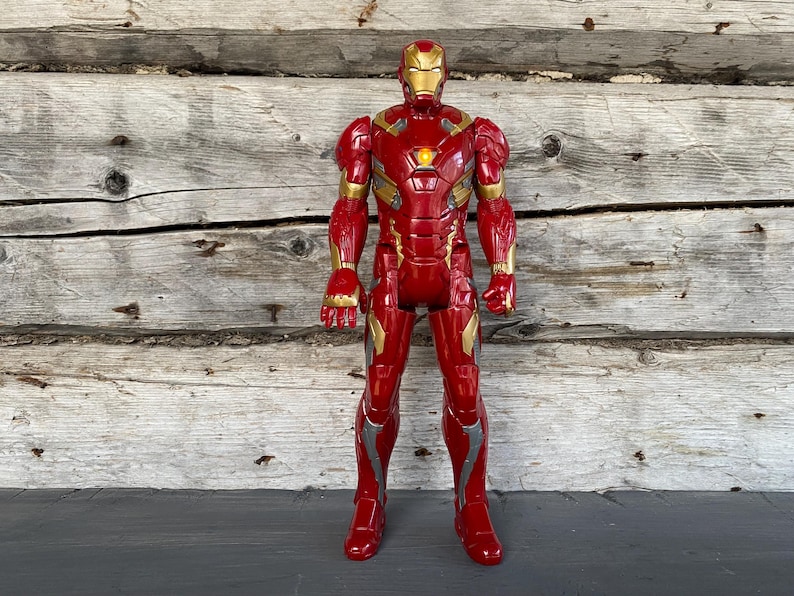 May include: A red and gold Iron Man action figure stands against a wooden background. The figure has a detailed design with gold accents on the arms, legs, and helmet. The figure is a collectible toy.