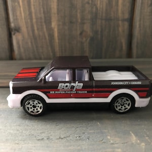 May include: A brown and red toy pickup truck with white accents. The truck has the text "Borla Exhaust" and "We Super Pickup Truck" on the side. The truck also has the text "Johnson City • Oxnard".