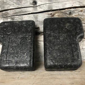Puede incluir: Dos insertos de embalaje de espuma negra moldeada con una superficie texturizada. Cada uno tiene una forma rectangular con una extensión curva en forma de asa. Los insertos tienen texto en relieve y detalles circulares. Descansan sobre una superficie de madera envejecida.