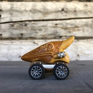 Könnte beinhalten: Ein Spielzeugauto mit einem braunen Dinosaurierkopf und -körper, montiert auf einem Fahrgestell mit vier silbernen Rädern. Der Dinosaurierkopf hat eine detaillierte Textur und ein rotes Auge. Das Auto steht vor einem verwitterten Holzhintergrund.