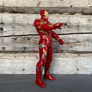 May include: A red and gold Iron Man action figure, standing with one arm extended. The figure has a gold helmet and accents, with silver details on the legs. The background is a weathered wooden wall.