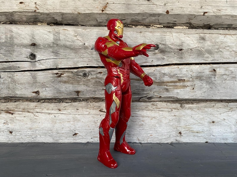 May include: A red and gold Iron Man action figure, standing with one arm extended. The figure has a gold helmet and accents, with silver details on the legs. The background is a weathered wooden wall.