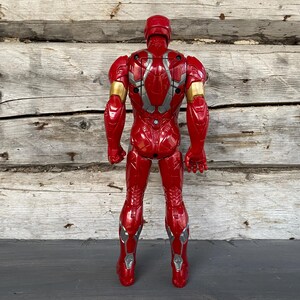 May include: A red Iron Man action figure, viewed from the back, with gold and silver accents. The toy is standing upright against a weathered wooden background. The figure has a helmet and detailed armor.