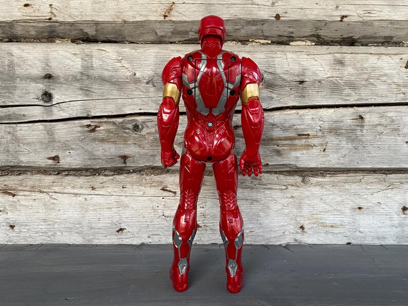 May include: A red Iron Man action figure, viewed from the back, with gold and silver accents. The toy is standing upright against a weathered wooden background. The figure has a helmet and detailed armor.