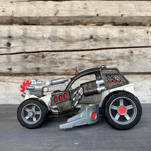 Puede incluir: Un coche de juguete blanco y gris con ruedas negras y detalles rojos. El coche tiene una estructura en forma de jaula y un arma adjunta en la parte delantera. Las ruedas tienen tapacubos rojos. El coche está sobre una superficie de madera.