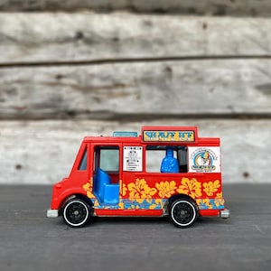May include: A red toy food truck with blue and yellow floral designs. The truck has black wheels and the words "SHAVE ICE" on top. The side of the truck has a logo and a list of items.
