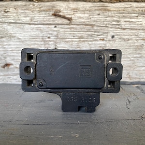 May include: A black rectangular electronic component with rounded corners, featuring a central raised section and two circular holes. The component has the numbers "038 9103" printed on it. The background is a weathered wooden surface.