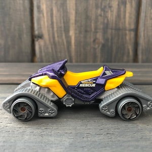 May include: A toy vehicle with a purple and yellow body, gray treads, and the word "RESCUE" printed on the side. The toy has two wheels and is designed to look like a rescue vehicle.