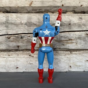 May include: A blue Captain America action figure with a white star on the back and a red, white, and blue design on the torso. The figure has red boots and gloves, and a brown belt. The right arm is raised in a salute.
