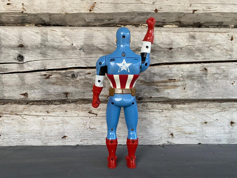 May include: A blue Captain America action figure with a white star on the back and a red, white, and blue design on the torso. The figure has red boots and gloves, and a brown belt. The right arm is raised in a salute.