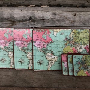May include: A set of vintage-style world map placemats and coasters. The placemats are rectangular, and the coasters are square. The maps feature a color palette of pink, green, and brown, with a compass rose design. The set is displayed on a wooden surface.