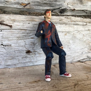 May include: A detailed action figure of a person in a dark blue pinstripe suit, splattered with red paint. The figure wears red and white sneakers. The background is a weathered wooden surface.
