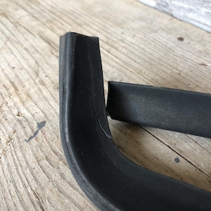 Op de afbeelding: Close-up van twee zwarte rubberen strips, één gebogen en de andere recht, op een verweerd houten oppervlak. De gebogen strip heeft een zichtbare scheur. De strips lijken deel uit te maken van een groter object of onderdeel.