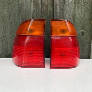 Könnte beinhalten: Ein Paar Fahrzeugrückleuchten mit geschwungenem Design, bestehend aus einem hellen, orangefarbenen Blinkerbereich im oberen Teil und einem größeren, horizontal gerippten roten Bremslichtbereich darunter, gefertigt aus durchscheinendem Kunststoff.