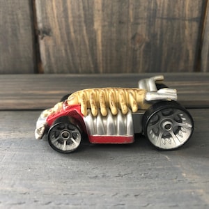 May include: A toy car with a unique design. The car features a red front, silver sides, and a tan engine detail. The wheels are black with silver rims. The car is a collectible toy.