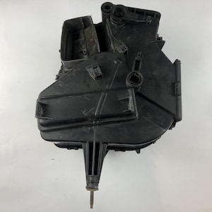 May include: A black plastic automotive part, possibly an air intake or filter housing, with various inlets, outlets, and mounting points. It has a rectangular shape with a central, elongated projection ending in a metal rod.