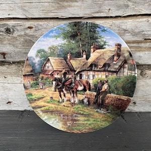 May include: A decorative plate featuring a pastoral scene with a horse-drawn cart, thatched-roof cottages, and figures near a pond. The plate has a blue border and is set against a rustic wooden background.