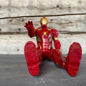 May include: A red and gold Iron Man action figure in a seated position, with outstretched hand. The figure has a detailed design, with a gold helmet and chest plate. The figure's feet are red, and the figure is on a gray surface.