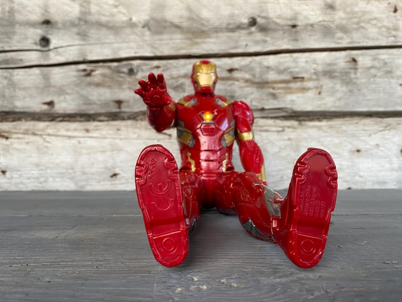 May include: A red and gold Iron Man action figure in a seated position, with outstretched hand. The figure has a detailed design, with a gold helmet and chest plate. The figure's feet are red, and the figure is on a gray surface.
