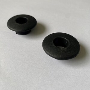 Op de afbeelding: Twee ronde zwarte rubberen doorvoertules met een glad, plat bovenoppervlak en een centraal gat. Elke tule heeft een licht verhoogde buitenrand en een cilindrische basis, ontworpen voor plaatsing in een gat ter bescherming.