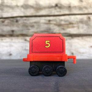 May include: A red toy train car with the number 5 in yellow. The car has black wheels and a red frame. The background is a wooden surface.