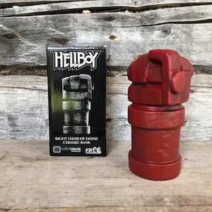 Puede incluir: Un banco de cerámica roja de la Mano Derecha de la Perdición de la franquicia Hellboy. El banco es una réplica detallada de la icónica mano del personaje. Una caja negra con el logotipo y texto de Hellboy está al lado.