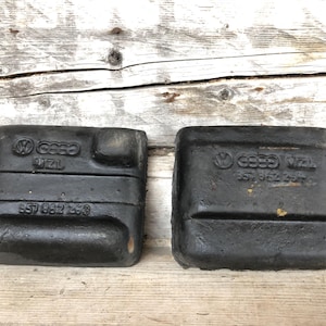 Puede incluir: Dos piezas automotrices negras y rectangulares con los logotipos de Volkswagen y Audi. Las piezas tienen las inscripciones "17ZL" y "857 862 253". Se encuentran sobre una superficie de madera envejecida.