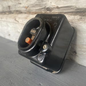 Può includere: Una presa elettrica nera con una porta aperta, che rivela i componenti interni. La presa ha una forma rettangolare con bordi arrotondati e una vite sul lato. Lo sfondo è una superficie di legno.