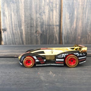 May include: A gold, black, and red toy race car with the text "SUPER GAMES" on the side. The car has red wheels with gold rims and a sleek, aerodynamic design. The car is set against a dark wood background.