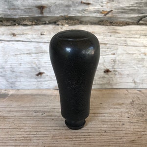 May include: A black, textured gear stick knob with a rounded top and a flared base. The knob is made of a solid material and has a slightly glossy finish. The background is a weathered wooden surface.