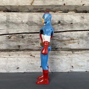 May include: A vintage Captain America action figure. The figure is primarily blue with red and white accents, including a red belt, gloves, and boots. The figure has a blonde head and a red stripe on top of the head.