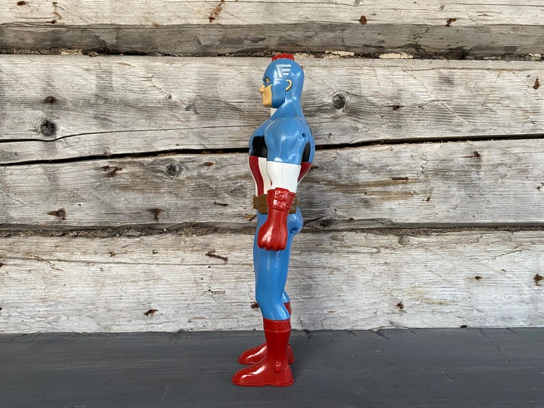 May include: A vintage Captain America action figure. The figure is primarily blue with red and white accents, including a red belt, gloves, and boots. The figure has a blonde head and a red stripe on top of the head.