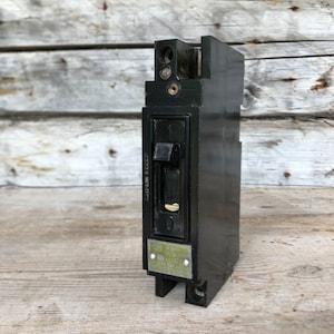 May include: A black electrical circuit breaker with a toggle switch and a small metal plate. The switch is in the 'off' position. The device has a rectangular shape and is made of a dark, possibly plastic material. The background is a weathered wooden surface.