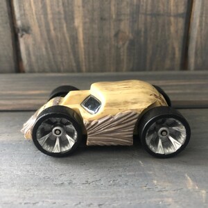 May include: A small, wooden toy car with a light tan body and black wheels. The car has a square window and detailed side panels. The wheels have silver-colored centers. The car is sitting on a gray surface.