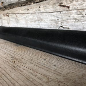 Könnte beinhalten: Ein langes, schwarzes, strukturiertes Objekt, möglicherweise eine Zierleiste, ist diagonal auf einer Holzoberfläche positioniert. Das Objekt hat eine leicht abgerundete Oberseite und eine gleichmäßige, dunkle Farbe. Der Holzuntergrund hat ein rustikales, verwittertes Aussehen.