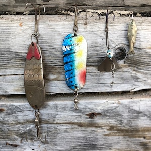 May include: A selection of vintage fishing lures displayed against a weathered wooden backdrop. The lures showcase diverse designs, including a fish-shaped lure, a blue and yellow lure, and a lure with a red top. Each lure is connected to a hook.