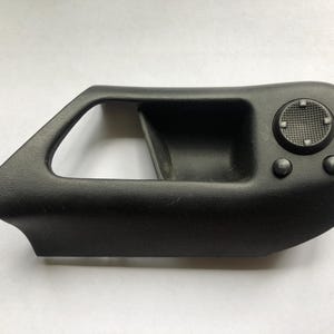 Puede incluir: Manija de puerta de coche de plástico negro con superficie texturizada. Cuenta con un área de agarre empotrada, un panel de control circular con un patrón de cruz y dos botones circulares más pequeños. El mango tiene forma triangular en un extremo.