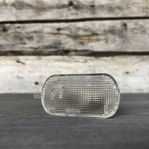 Puede incluir: Una luz de posición lateral automotriz clara y rectangular con una superficie texturizada. La luz tiene bordes redondeados y está colocada horizontalmente. El fondo es una superficie de madera desgastada.