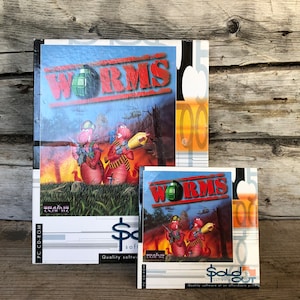 Puede incluir: Se muestran dos cajas de juegos PC-CD-ROM vintage de "Worms". La caja más grande presenta el título del juego en rojo, una granada verde y gusanos de dibujos animados en una escena de campo de batalla. La caja más pequeña refleja el diseño. Las cajas están sobre una superficie de madera.