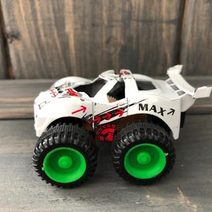 May include: A white toy truck with black tires and green rims. The truck has red and black graphics and the word "MAX" printed on the side. The toy is designed to look like a monster truck.