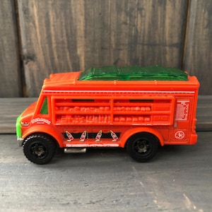 May include: A toy food truck in orange with green accents. The truck has black wheels and a green roof. The side of the truck has a graphic of bottles and the text "Sabores".