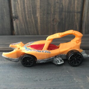 May include: Orange toy car with a red interior and black wheels. The car has a unique design with a curved roll bar and decorative blue accents. The car is a toy vehicle.