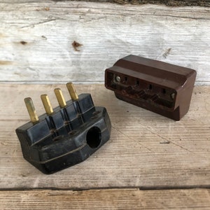 May include: Two vintage electrical plugs. One is black with brass pins, the other is brown. The plugs are made of a hard plastic material and are designed to connect to electrical sockets. The plugs are set against a weathered wooden background.
