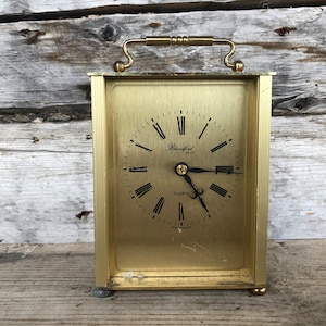 Puede incluir: Un reloj de cuarzo rectangular dorado con asa. La esfera del reloj tiene números romanos y manecillas negras, con la marca "Blanford 1973" impresa en la esfera. El reloj está sobre una superficie de madera.
