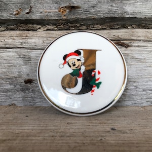 Op de afbeelding: Een witte, ronde decoratieve plaat met een gouden rand. De plaat heeft een gouden letter "J" met Mickey Mouse met een kerstmuts en een zuurstok. Het ontwerp is feestelijk en heeft een vakantiethema.