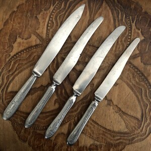 May include: Four vintage silver knives with ornate handles are arranged diagonally on a wooden surface. The blades are slightly curved and show signs of wear. The wooden surface has a carved design, adding to the antique aesthetic.