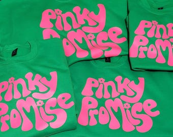 May include: A collection of bright green crewneck sweatshirts featuring the text Pinky Promise printed in a bold, vibrant pink font. Each sweatshirt displays the same graphic design with a small flower icon integrated into the lettering.