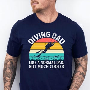 May include: Navy blue t-shirt with a colorful graphic featuring a diver silhouette against a sunset design. The text "DIVING DAD LIKE A NORMAL DAD, BUT MUCH COOLER" is printed in white. The shirt is short-sleeved.